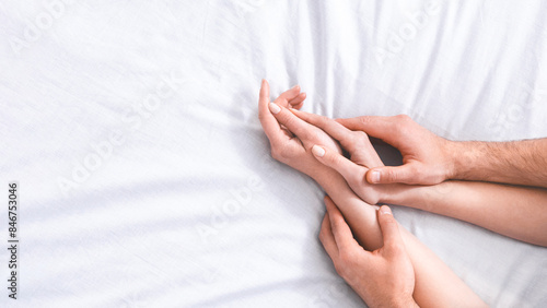 This image shows the hands of a couple intertwined on a white bed, suggesting intimacy and connection, cropped, copy space