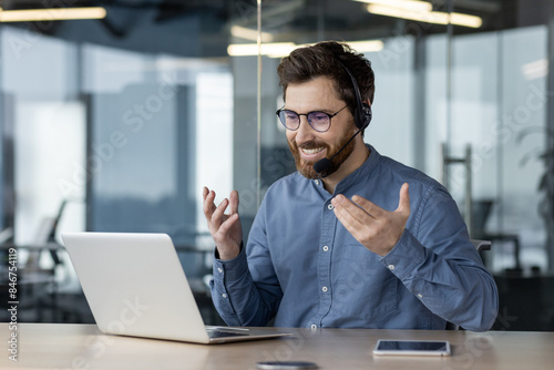 Smiling young man talking on a video call on a laptop, sitting in the office in a headset and gesturing with his hands to explain