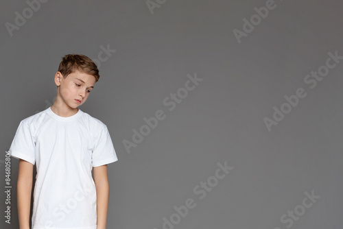 A sad teenage boy gazing downwards, expressing despondency He stands isolated with a grey background, copy space