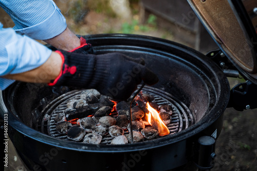 A man is safely lighting charcoal on a grill for an outdoor BBQ preparation, wearing safety gloves. The keywords include BBQ, charcoal, fire, cooking, and grilling