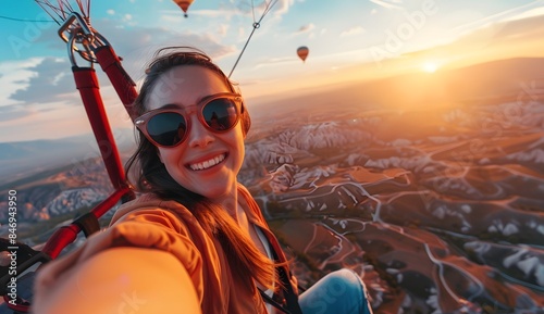 Happy smiling woman in sunglasses taking a selfie photo while sitting on a hot air balloon, flying above the valley at sunrise