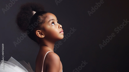 Young African American girl in ballet tutu looking upwards against dark background. Image conveys innocence, hope, and aspiration with focus on her expressive profile