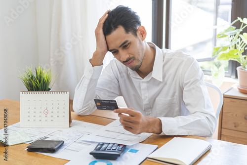 Financial concept, owe asian young male sitting suffer, stressed and confused by calculate expense from invoice, holding mock up credit card, no money to pay mortgage or loan. Debt, bankrupt people.