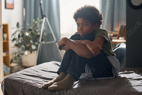 Full length portrait of upset young African American boy sitting on bed alone and hugging knees copy space