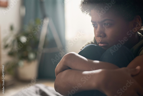 Dramatic close up portrait of Black boy crying and hugging knees in hazy dark room with blurred background copy space