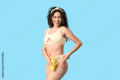 Beautiful young African-American woman in swimsuit pointing at heart made of sunscreen cream on her leg against blue background