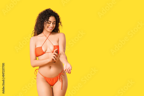 Beautiful young African-American woman in swimsuit applying sunscreen cream on her hand against yellow background
