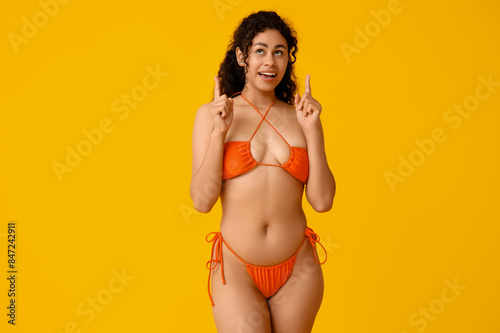 Beautiful young African-American woman in stylish orange swimsuit pointing at something on yellow background