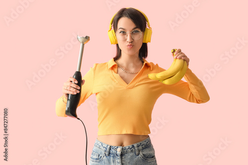 Young woman in headphones with hand blender and banana blowing kiss on pink background
