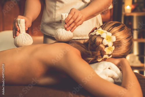 Closeup hot herbal ball spa massage body treatment, masseur gently compress herb bag on woman body. Tranquil and serenity of aromatherapy recreation in warm lighting of candles at spa salon. Quiescent