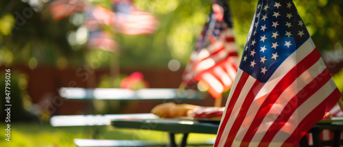 An outdoor setting features American flags waving in the gentle breeze, surrounded by greenery and picnic tables, evoking a patriotic atmosphere.