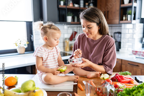 The mother prepared a healthy fruit and yogurt meal rich in vitamins, and she's feeding her cute little baby daughter at the home kitchen counter