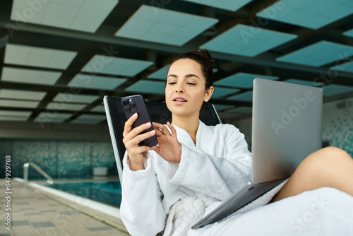 A young, beautiful brunette woman relaxes in a bathrobe by an indoor spa pool, focused on her cell phone.