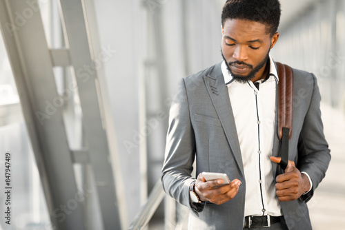 A business traveler black man is standing outside a modern airport, using his phone. The environment is sleek and professional, emphasizing the blend of work and travel.