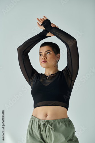 A young woman with short hair poses in a stylish black top and green pants on a grey background.