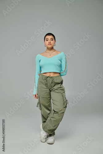 A stylish young woman with short hair posing confidently in a trendy crop top and cargo pants against a grey backdrop.