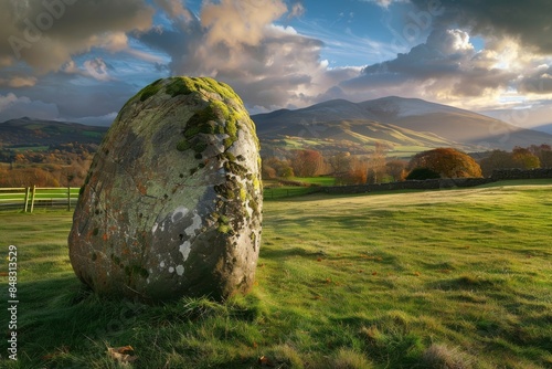 A landscape photograph of the English Lake District, in an open valley. A large boulder covered with moss and lichen is in the foreground, background are rolling mountains under a dramatic sky.