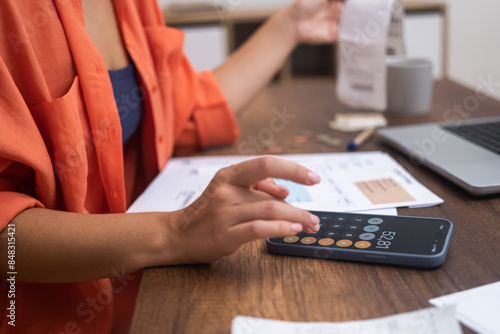 Budget Control: At her desk, alongside a laptop, a woman manages household finances using her smartphone calculator