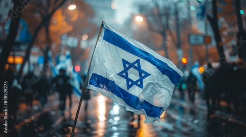 The Israeli flag is prominently displayed waving amidst a rainy urban street setting, symbolizing patriotism