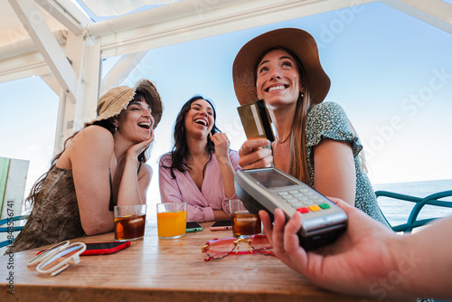 Happy young group of only women paying the bill with a contactless credit card in cocktail beach bar. Female smiling holding a creditcard and giving a payment transaction to the cashier on vacations