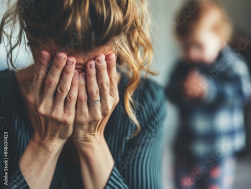 Stressed and frustrated young mother hiding face in hands with toddler child in blurry background - ai