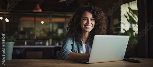 Female freelancer at a wooden table smiling with hand on chin gazes at the camera while working on a new project on her laptop with copy space image