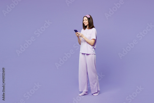 Full body young calm woman wear pyjamas jam sleep eye mask rest relax at home hold in hand use mobile cell phone isolated on plain pastel light purple background studio. Good mood night nap concept.