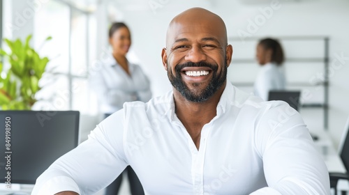 In an office with coworkers, a confident young mixed-race businessman smiles with hand on chin. Determined leader and entrepreneur willing to succeed in business with team