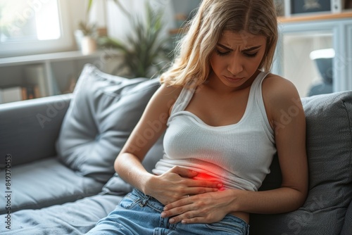 Young woman sitting on a couch, holding her stomach in pain, highlighting abdominal discomfort or menstrual cramps with a red glow.