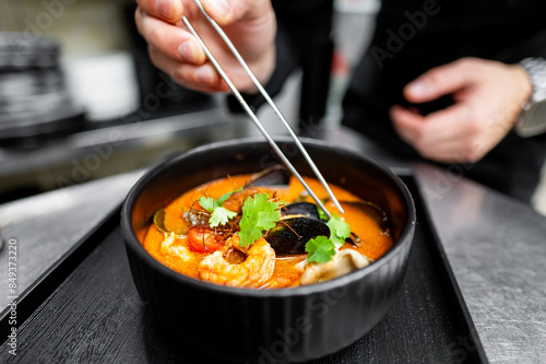 Close-up of a chef’s hands garnishing a gourmet seafood soup with fresh herbs in a black bowl, showcasing the art of fine dining