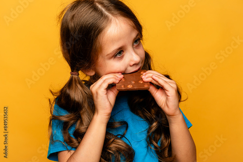 Cute little girl eating chocolate bars with nuts on yellow background, closeup. The concept of children's happiness and love for sweets