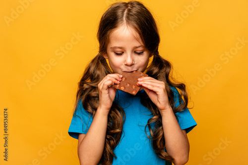 Cute little girl eating chocolate bars with nuts on yellow background, closeup. The concept of children's happiness and love for sweets