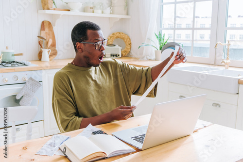 African American man sitting in bright kitchen, looking shocked while holding long receipt. He has laptop, notebook, and receipts on table, indicating he reviewing expenses or budgeting.