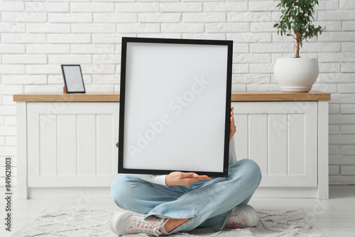 Young woman sitting near chest of drawers and holding blank picture frame in room