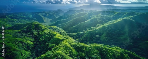 Aerial view of lush green mountains with sun rays illuminating the landscape and a dramatic cloudy sky overhead, creating a picturesque scene.
