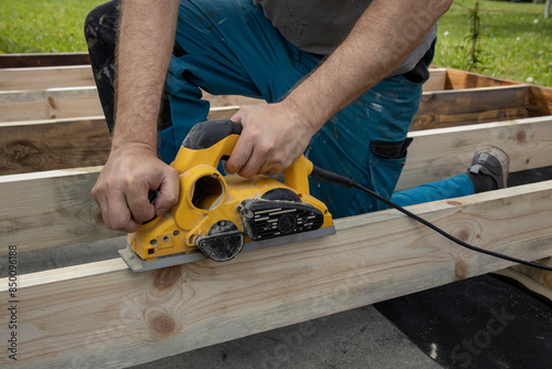 A carpenters hands are shown using a power planer to smooth out a wooden beam. The carpenter is wearing blue pants and is kneeling on the wooden beams.