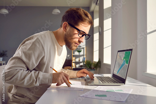 Portrait of professional cartographer looking at laptop monitor screen, working with printed cadastral map at table on workplace. Young man analyzing cadastral map searching for building plot.