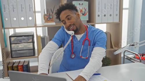 Handsome african american man with a beard working in a veterinary clinic, wearing scrubs and a stethoscope, multitasking with a phone and computer in a bright, organized interior.