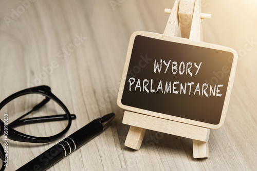 Black writing board on a wooden frame with the inscription "Wybory Parlamentarne", next to black glasses and a pen (selective focus) translation: Parliamentary Elections