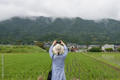 里山の田園風景をスマートフォンで撮影する女性の後ろ姿