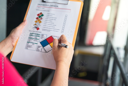 Action of a safety supervisor hand is checking on checklist of chemical and hazardous material form to verify the risk with blurred background of factory storage workplace. Industrial working scene.