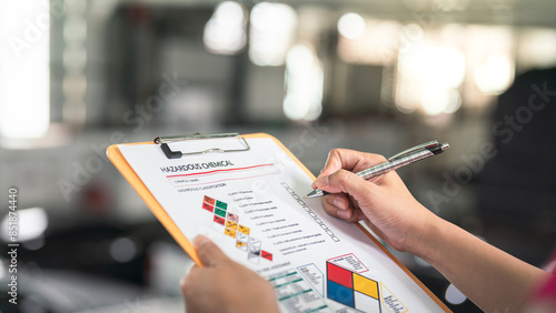 Action of a safety supervisor hand is checking on checklist of chemical and hazardous material form to verify the risk with blurred background of factory storage workplace. Industrial working scene.