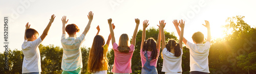 Happy children outdoors in summer raise their hands together, reaching for sky. Rear view of teenage boys and girls in casual clothes standing in row with raised arms in sunlight. Panoramic web banner