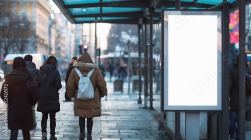 People walk down a city street near a bus stop with a blank advertisement display during a cold day.