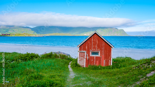 A weathered red cabin sits on a white sand beach in Norway, with a turquoise sea and a green mountain range in the distance. Ramberg Beach Lofoten Norway