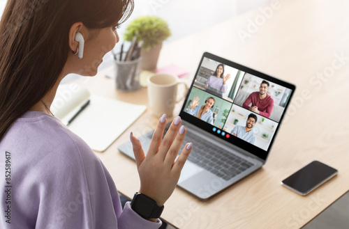 This image shows a young woman participating in a video conference meeting at home. She is wearing a purple sweatshirt and has her hand raised to the camera, as if she is about to speak.