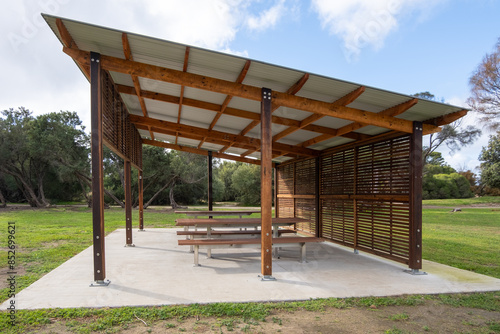 A wooden gazebo or pavilion with a roof, featuring public bench seats, serves as a covered picnic area in a park, offering ventilation and recreational rest facilities.