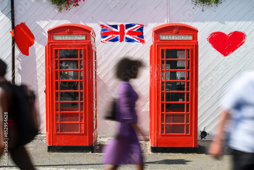 Classic red British telephone booths with blurred pedestrians in motion. The iconic red telephone boxes are set against a white wall with a painted Union Jack and hearts.