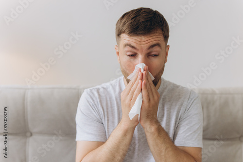 Unhappy middle aged man suffering from fever and flu, blowing nose in napkin, sitting in living room interior. Covid-19 lockdown, treatment of illness, cold and runny