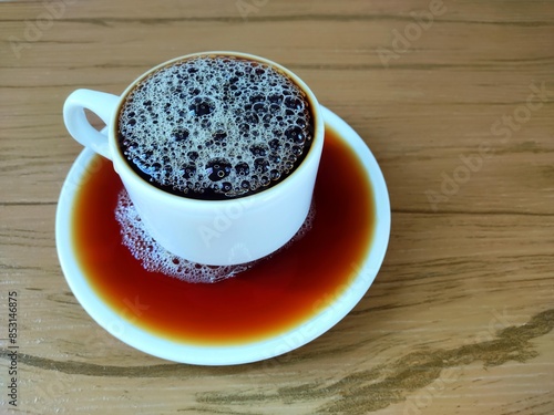 Full cup of coffee with foam, which overflowed and fell on top of the pyrex dish on the wooden table. 